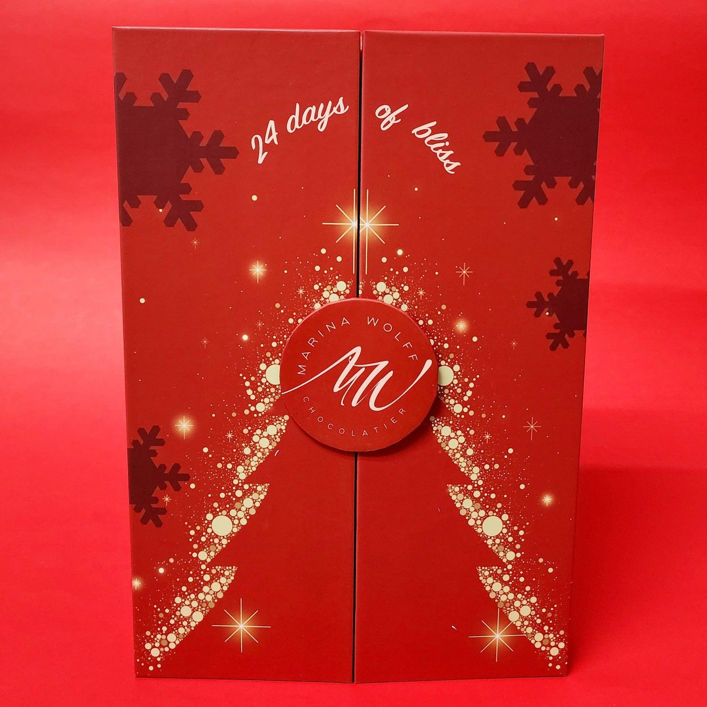 A red rectangular box with a silhouette of a tree surrounded by snowflakes and stars written ' 24 days of bliss' on top of it on a red background.